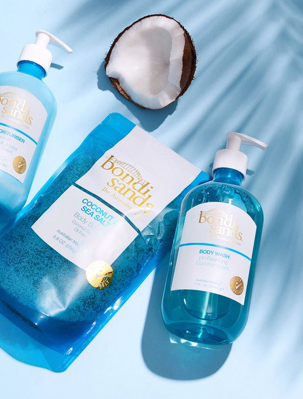 Flat lay of the BondiSandsEU Coconut Body Bundle—body wash, scrub, and moisturiser in blue packaging—beside a halved coconut on a light blue background with soft shadows.
