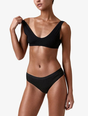 A woman in a matching black ribbed bra and panties set stands against a white background, showing off her glowing skin from a BondiSandsEU Salon Solution Dark spray tan. She lifts a bra strap with her left hand; her head is partially out of frame.