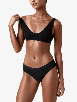 A woman in a black bikini poses against a white background, lifting one top strap. Her skin glows with a flawless tan, suggesting use of BondiSandsEU Salon Solution Dark Duo and its subtle coconut scent. Her face is partially out of frame.